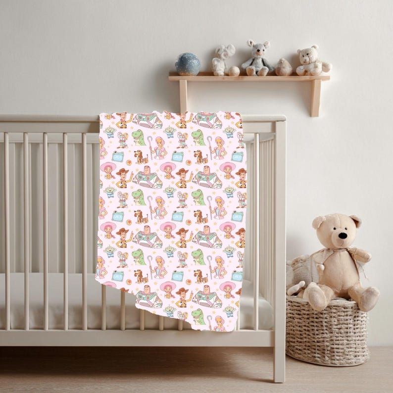 May include: A baby blanket featuring a repeating pattern of cartoon characters in various colours, including green, pink, and yellow. The blanket is draped over a white cot. A teddy bear sits in a wicker basket.
