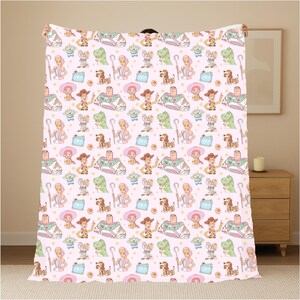 May include: A pink blanket with a repeating pattern of cartoon characters, including a cowboy, space ranger, and dinosaur. The blanket has a soft, plush appearance and is likely made of a comfortable material. The design includes stars and other elements.