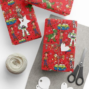May include: Red wrapping paper featuring a repeating Toy Story character pattern with Woody, Buzz Lightyear, Rex, and the Aliens wearing Santa hats, accented by Christmas lights and snowflakes. Includes twine, white gift tags, and black scissors.