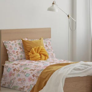 May include: A twin bed with a light wood headboard and frame, featuring a light pink duvet cover with a repeating pattern of cartoon characters, a mustard yellow square pillow, a mustard yellow knot pillow, and a white throw blanket.