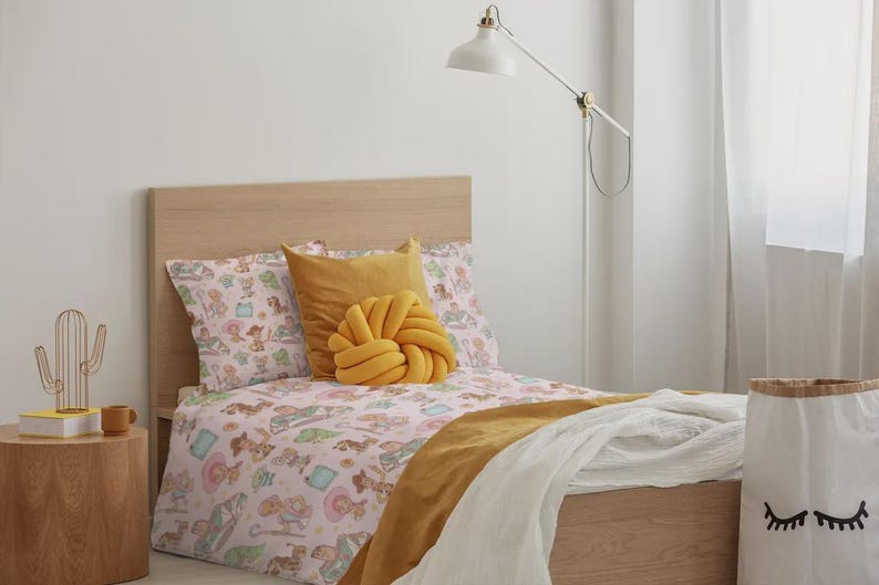 May include: A twin bed with a light wood headboard and frame, featuring a light pink duvet cover with a repeating pattern of cartoon characters, a mustard yellow square pillow, a mustard yellow knot pillow, and a white throw blanket.