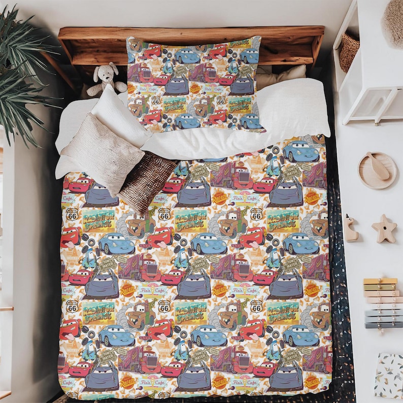 Pode incluir: Conjunto de roupa de cama branco com um padr&atilde;o repetido de carros de desenho animado, sinais da Rota 66 e gr&aacute;ficos relacionados. O design apresenta carros em azul, vermelho e cinza, criando uma est&eacute;tica vibrante e l&uacute;dica.