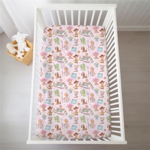 May include: A crib sheet featuring a repeating pattern of Toy Story characters including Woody, Buzz Lightyear, Bo Peep, Rex, Slinky Dog, and the green aliens on a light pink background with small yellow stars scattered throughout the design.