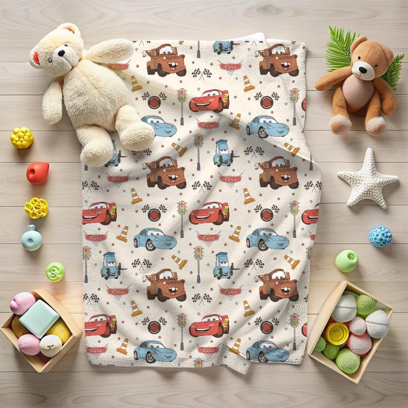 May include: A cream-coloured fleece blanket featuring a repeating pattern of red race cars, brown tow trucks, blue sports cars, traffic lights, orange traffic cones, and checkered flags, surrounded by two teddy bears and small toys.