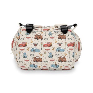 Puede incluir: Una mochila de color crema con un patr&oacute;n repetitivo de personajes de dibujos animados de la pel&iacute;cula Cars, incluyendo coches de carreras rojos, gr&uacute;as marrones y coches azules, junto a sem&aacute;foros, conos naranjas y banderas a cuadros.