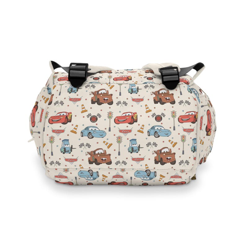 Puede incluir: Una mochila de color crema con un patr&oacute;n repetitivo de personajes de dibujos animados de la pel&iacute;cula Cars, incluyendo coches de carreras rojos, gr&uacute;as marrones y coches azules, junto a sem&aacute;foros, conos naranjas y banderas a cuadros.