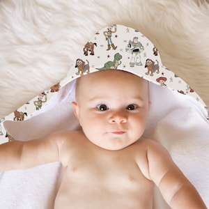 May include: A baby wearing a hooded towel featuring a pattern of cartoon characters. The towel's hood displays images of Woody, Buzz Lightyear, Rex, and other characters from the Toy Story franchise, set against a cream background with star accents.