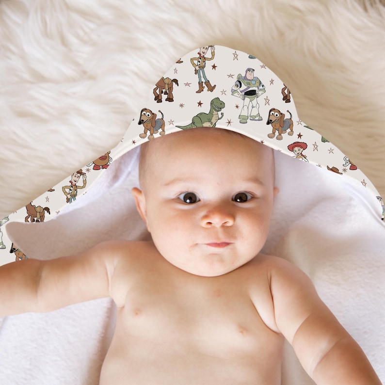 May include: A baby wearing a hooded towel featuring a pattern of cartoon characters. The towel's hood displays images of Woody, Buzz Lightyear, Rex, and other characters from the Toy Story franchise, set against a cream background with star accents.