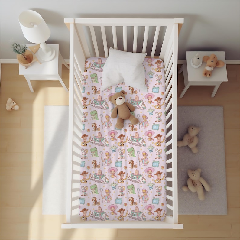 May include: A white wooden crib featuring a fitted sheet with a repeating pattern of cartoon characters including a space ranger, a cowboy, a cowgirl, a green dinosaur, and a three-eyed alien. A white pillow and a brown teddy bear sit on the sheet.