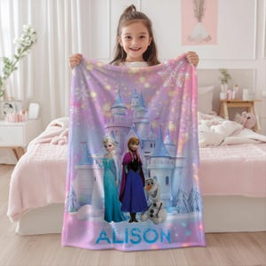 May include: A pink and purple blanket featuring characters from the movie Frozen, including Elsa, Anna, and Olaf, in front of a castle. The blanket has the name "Alison" in blue at the bottom. Snowflakes and glittery effects are also present.
