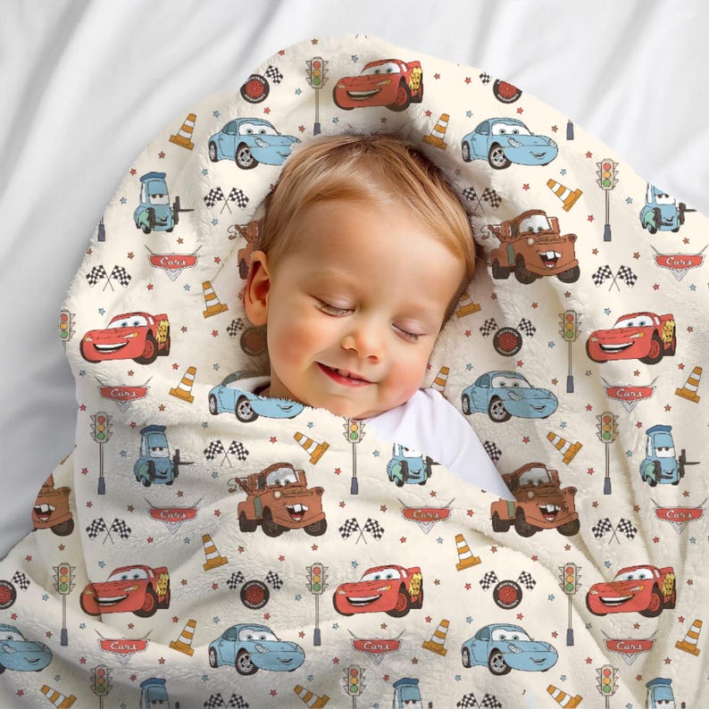 May include: A soft, cream-coloured fleece blanket featuring a repeating pattern of characters from the movie Cars, including red race cars, blue sports cars, and brown tow trucks, interspersed with traffic lights, orange cones, and chequered flags.
