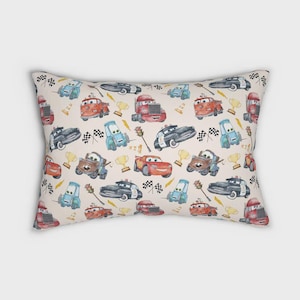 Op de afbeelding: Een rechthoekig sierkussen met een lichtbeige achtergrond met een herhalend patroon van cartoonraceauto's, politieauto's, takelwagens, gouden trofeeën, geblokte vlaggen, verkeerslichten en gele bliksemschichten in aquarel-stijl.