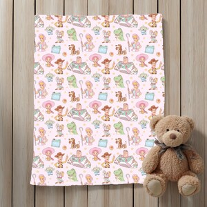 May include: A pink blanket with a repeating pattern of cartoon characters, including a cowboy, space ranger, and dinosaur. A brown teddy bear with a grey ribbon sits to the right of the blanket. The blanket is on a wooden surface.