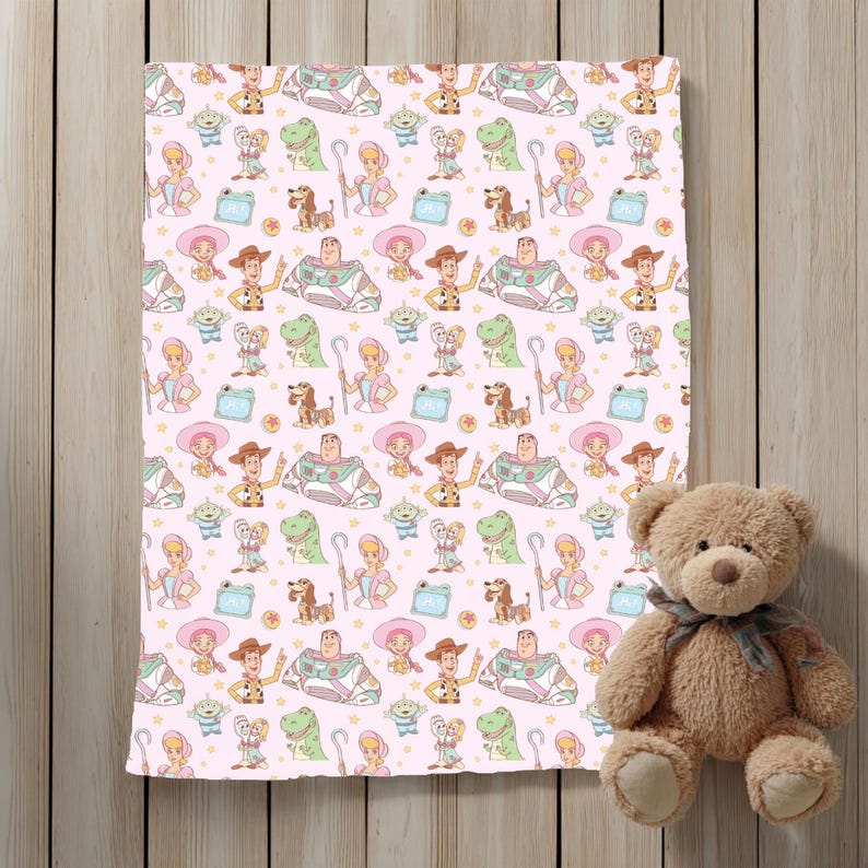 May include: A pink blanket with a repeating pattern of cartoon characters, including a cowboy, space ranger, and dinosaur. A brown teddy bear with a grey ribbon sits to the right of the blanket. The blanket is on a wooden surface.
