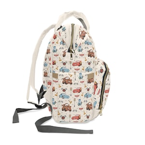 Puede incluir: Una mochila de color crema con un estampado de coches de dibujos animados, conos de tr&aacute;fico y banderas de cuadros. La bolsa incluye un asa superior, correas grises ajustables para los hombros, un bolsillo frontal y cremalleras doradas con tiradores.