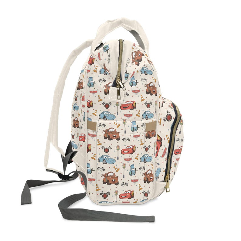 Puede incluir: Una mochila de color crema con un estampado de coches de dibujos animados, conos de tr&aacute;fico y banderas de cuadros. La bolsa incluye un asa superior, correas grises ajustables para los hombros, un bolsillo frontal y cremalleras doradas con tiradores.