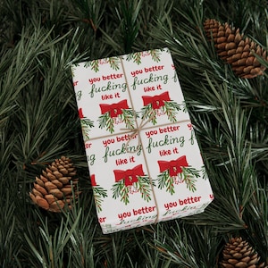 May include: A rectangular gift wrapped in white paper featuring a repeating pattern of red bows, green mistletoe sprigs, and the red and green text "you better fucking like it" tied with thin brown twine.