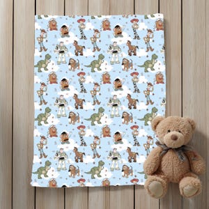 K&ouml;nnte beinhalten: Eine hellblaue Decke mit einem sich wiederholenden Muster aus Toy Story-Figuren wie Woody, Buzz Lightyear, Jessie, Rex, Mr. Kartoffelkopf, Slinky Dog und Bullseye, durchsetzt mit wei&szlig;en Wolken und kleinen Sternen, neben einem braunen Teddyb&auml;ren.
