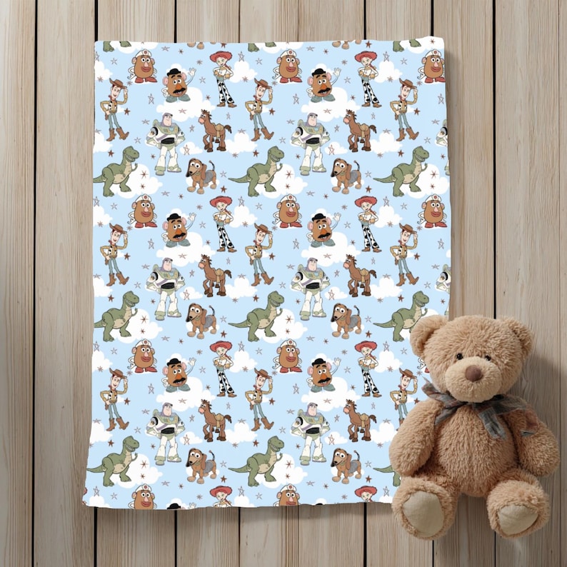 K&ouml;nnte beinhalten: Eine hellblaue Decke mit einem sich wiederholenden Muster aus Toy Story-Figuren wie Woody, Buzz Lightyear, Jessie, Rex, Mr. Kartoffelkopf, Slinky Dog und Bullseye, durchsetzt mit wei&szlig;en Wolken und kleinen Sternen, neben einem braunen Teddyb&auml;ren.