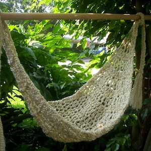 Puede incluir: Una hamaca de crochet blanca colgada de una barra de madera. La hamaca está hecha de un hilo grueso y suave y tiene un tejido suelto y abierto. La hamaca está colgada en un jardín con follaje verde en el fondo.