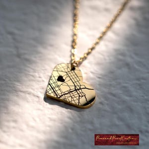May include: A gold-toned heart-shaped pendant necklace featuring an engraved black street map design with a small heart cutout. The pendant hangs from a delicate gold-toned chain, reflecting light across its polished surface.