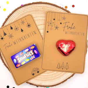 May include: Two kraft paper cards with the text "Frohe Weihnachten" and festive illustrations. One card has a Smarties Mini box, the other a red heart-shaped chocolate. The cards are on a wooden slice.