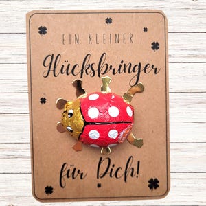 Könnte beinhalten: Ein rot-weißes Schokoladenbonbon in Form eines Marienkäfers auf einer braunen Karte. Die Karte hat deutschen Text und Kleeblatt-Illustrationen. Der Marienkäfer hat goldfarbene Flügel und Beine.