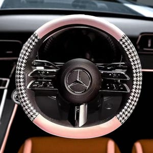 May include: A close-up of a car steering wheel. The wheel features a black and white houndstooth pattern on the sides, with a light pink band and a black center. The center has a silver emblem.