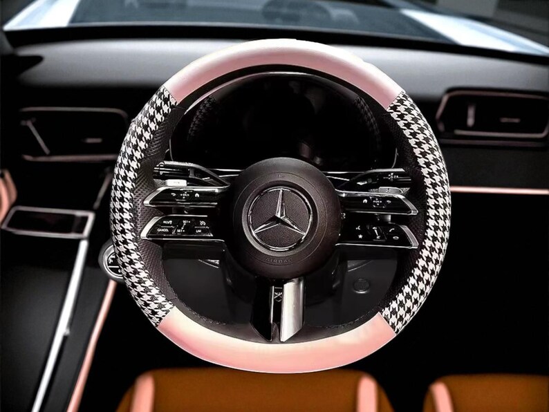 May include: A close-up of a car steering wheel. The wheel features a black and white houndstooth pattern on the sides, with a light pink band and a black center. The center has a silver emblem.