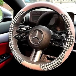 May include: A car steering wheel with a black and white houndstooth pattern on the sides and a light pink top and bottom. The center of the wheel features the Mercedes-Benz logo. The interior of the car is visible, with red and black accents.