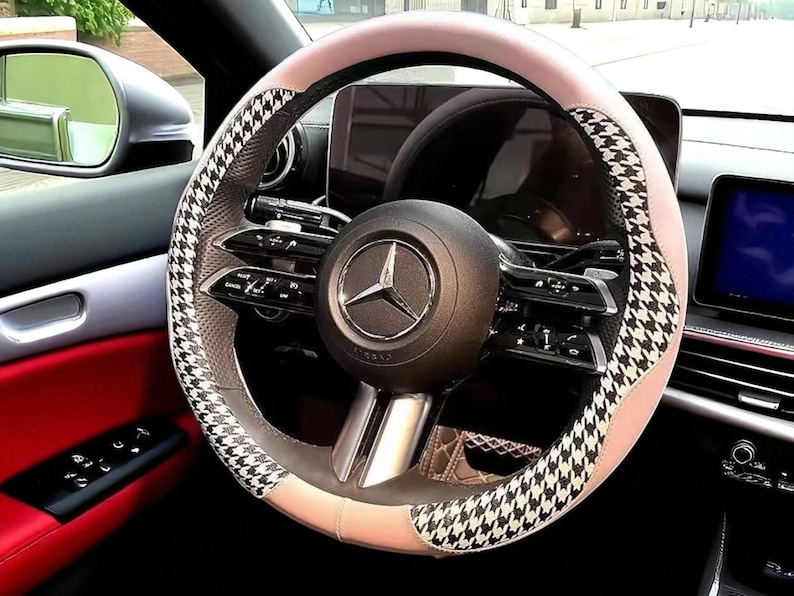 May include: A car steering wheel with a black and white houndstooth pattern on the sides and a light pink top and bottom. The center of the wheel features the Mercedes-Benz logo. The interior of the car is visible, with red and black accents.
