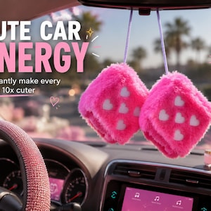 Op de afbeelding: Twee pluizige roze dobbelstenen auto-accessoires met witte hartpatronen hangen aan de achteruitkijkspiegel van een auto. Het interieur van de auto heeft een met strass versierd stuurwiel en een digitaal display. De tekst luidt "CUTE CAR ENERGY".