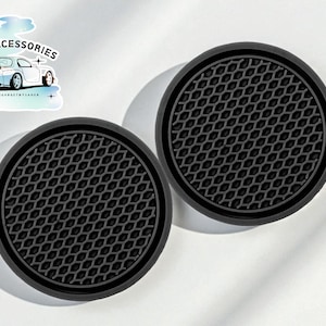 May include: Two black car coasters with a textured, honeycomb pattern. The coasters are round with a raised edge. A sticker in the upper left corner reads "CAR ACCESSORIES" with a car graphic.