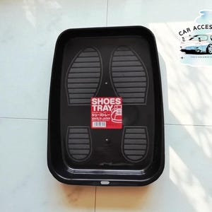 Puede incluir: Bandeja rectangular negra para zapatos con superficie texturizada. La bandeja tiene un diseño de huellas y las palabras "SHOES TRAY" en blanco y rojo. Una pegatina con las palabras "CAR ACCESSORIES" y un gráfico de coche está en la esquina superior derecha.
