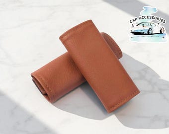 Può includere: Due copricintura di sicurezza per auto in pelle marrone sono esposti su una superficie di marmo bianco. Le coperture sono rettangolari con bordi arrotondati e sembrano essere realizzate in un materiale liscio e strutturato. Un adesivo con il testo "Car Accessories" e una grafica di auto è sullo sfondo.