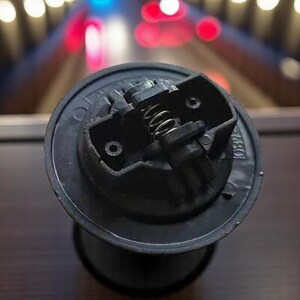 May include: A close-up of a black metal bottle stopper with a spring mechanism. The stopper has a circular shape and a textured surface.