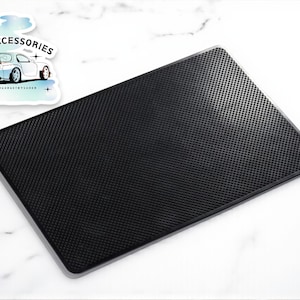 Puede incluir: Una alfombrilla negra rectangular para accesorios de coche con una superficie texturizada y punteada. La alfombrilla tiene un borde gris y se muestra sobre una superficie blanca. Una pegatina con el texto "CAR ACCESSORIES" y un gráfico de coche está en la esquina superior izquierda.