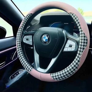 May include: A car steering wheel with a pink and black and white houndstooth pattern cover. The steering wheel has a BMW logo in the center. The car interior is black.