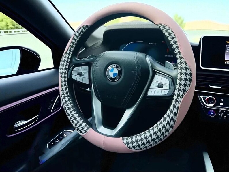 May include: A car steering wheel with a pink and black and white houndstooth pattern cover. The steering wheel has a BMW logo in the center. The car interior is black.