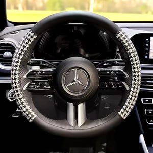 May include: Black leather steering wheel with a houndstooth pattern on the sides. The center features the Mercedes-Benz logo. The dashboard and other car components are visible in the background.