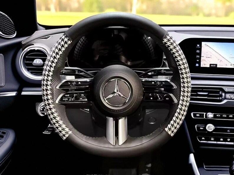 May include: Black leather steering wheel with a houndstooth pattern on the sides. The center features the Mercedes-Benz logo. The dashboard and other car components are visible in the background.