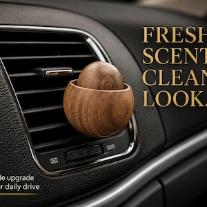 Puede incluir: Un ambientador de coche esférico de madera fijado a una rejilla de ventilación negra. El texto "FRESH SCENT. CLEAN LOOK." está impreso en dorado a la derecha, y "A simple upgrade for your daily drive" está impreso en blanco abajo a la izquierda.