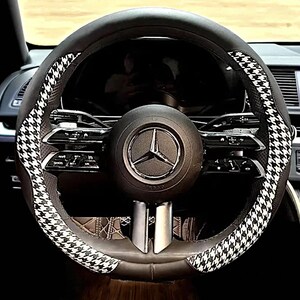 May include: Black leather steering wheel cover with a houndstooth pattern in black and white. The steering wheel has a Mercedes-Benz logo in the center. The interior of the car is visible in the background.
