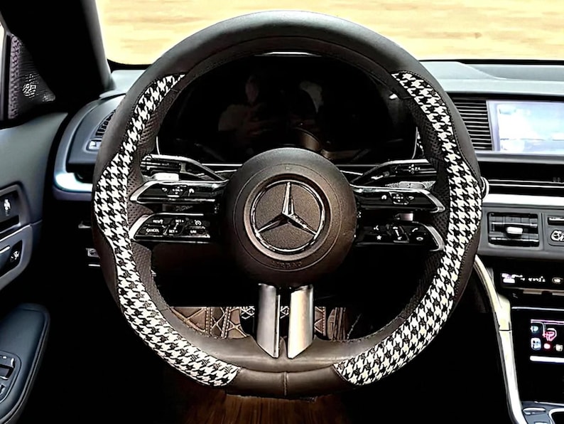 May include: Black leather steering wheel cover with a houndstooth pattern in black and white. The steering wheel has a Mercedes-Benz logo in the center. The interior of the car is visible in the background.
