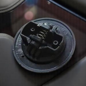 May include: Close-up of a black plastic gas cap with a spring-loaded mechanism. The cap has a circular shape and a textured surface.