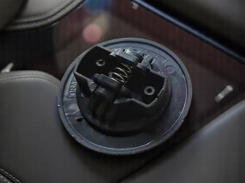 May include: Close-up of a black plastic gas cap with a spring-loaded mechanism. The cap has a circular shape and a textured surface.