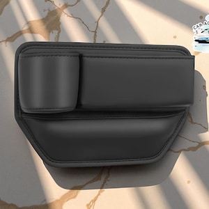 May include: Black car seat organizer with a cup holder and storage compartment. The organizer is made of a smooth, matte material and features a stitched design. The product has the text "CAR ACCESSORIES" on a sticker in the upper right corner.
