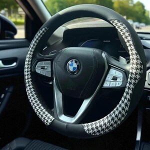 May include: Black and white houndstooth pattern steering wheel cover on a car's steering wheel. The cover is black with a white houndstooth pattern on the top and sides. The car's interior is black.