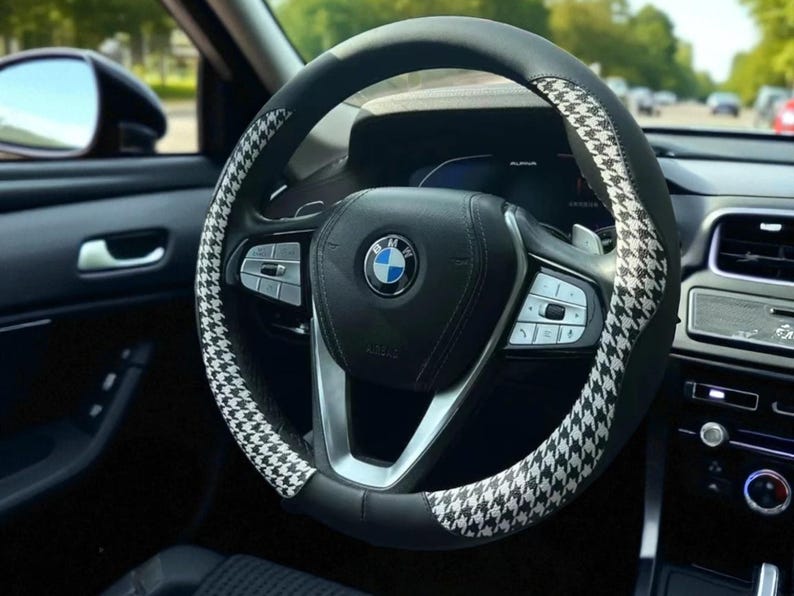 May include: Black and white houndstooth pattern steering wheel cover on a car's steering wheel. The cover is black with a white houndstooth pattern on the top and sides. The car's interior is black.