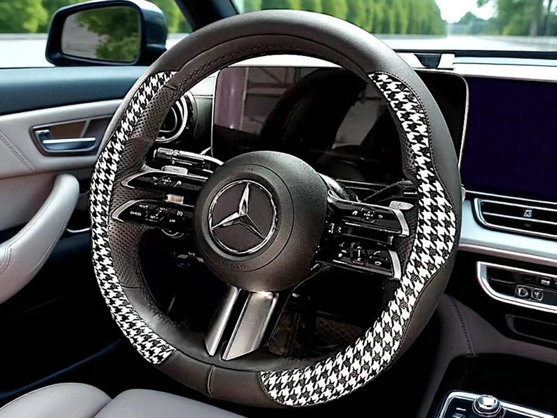 May include: A black and gray steering wheel cover with a houndstooth pattern. The cover is designed to fit a Mercedes-Benz vehicle. The steering wheel has a black center with the Mercedes-Benz logo.
